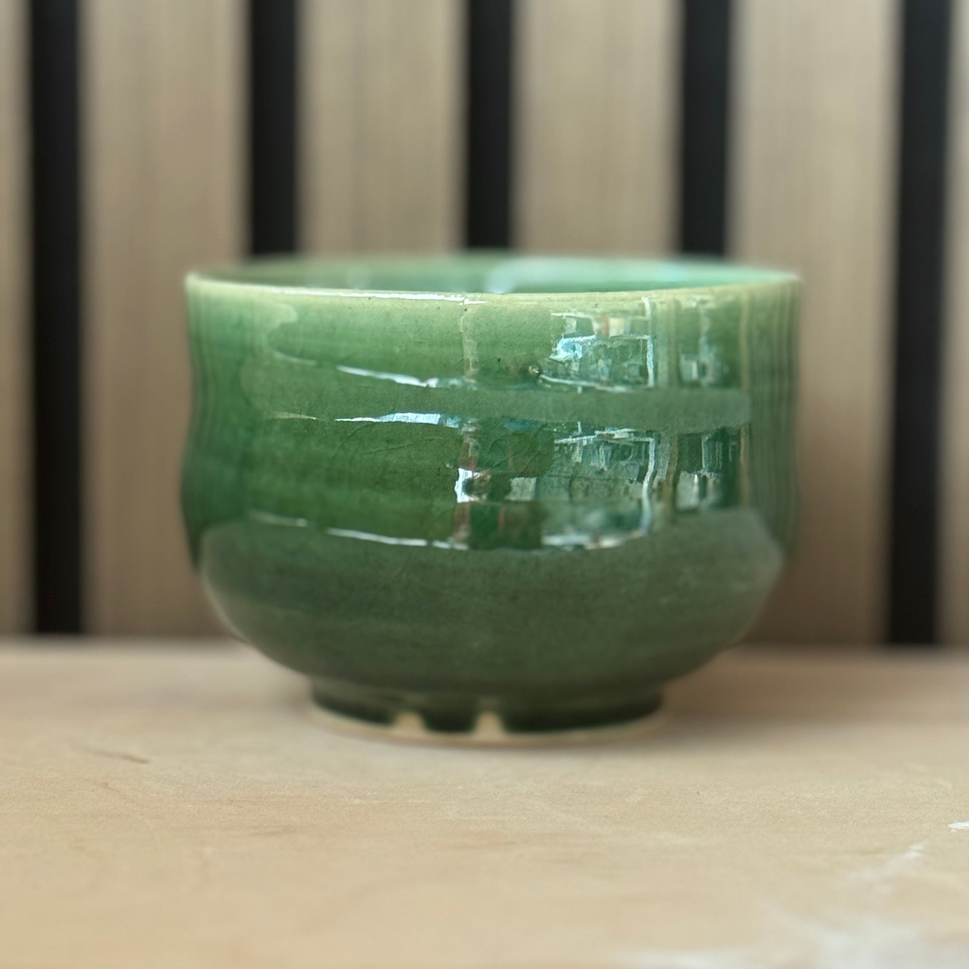 A glossy green ceramic cup with a wide rim and small base, shown on a light wooden surface with a blurred striped background.