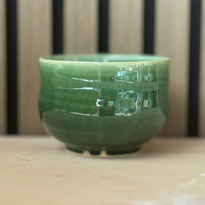 A glossy green ceramic cup with a wide rim and small base, shown on a light wooden surface with a blurred striped background.