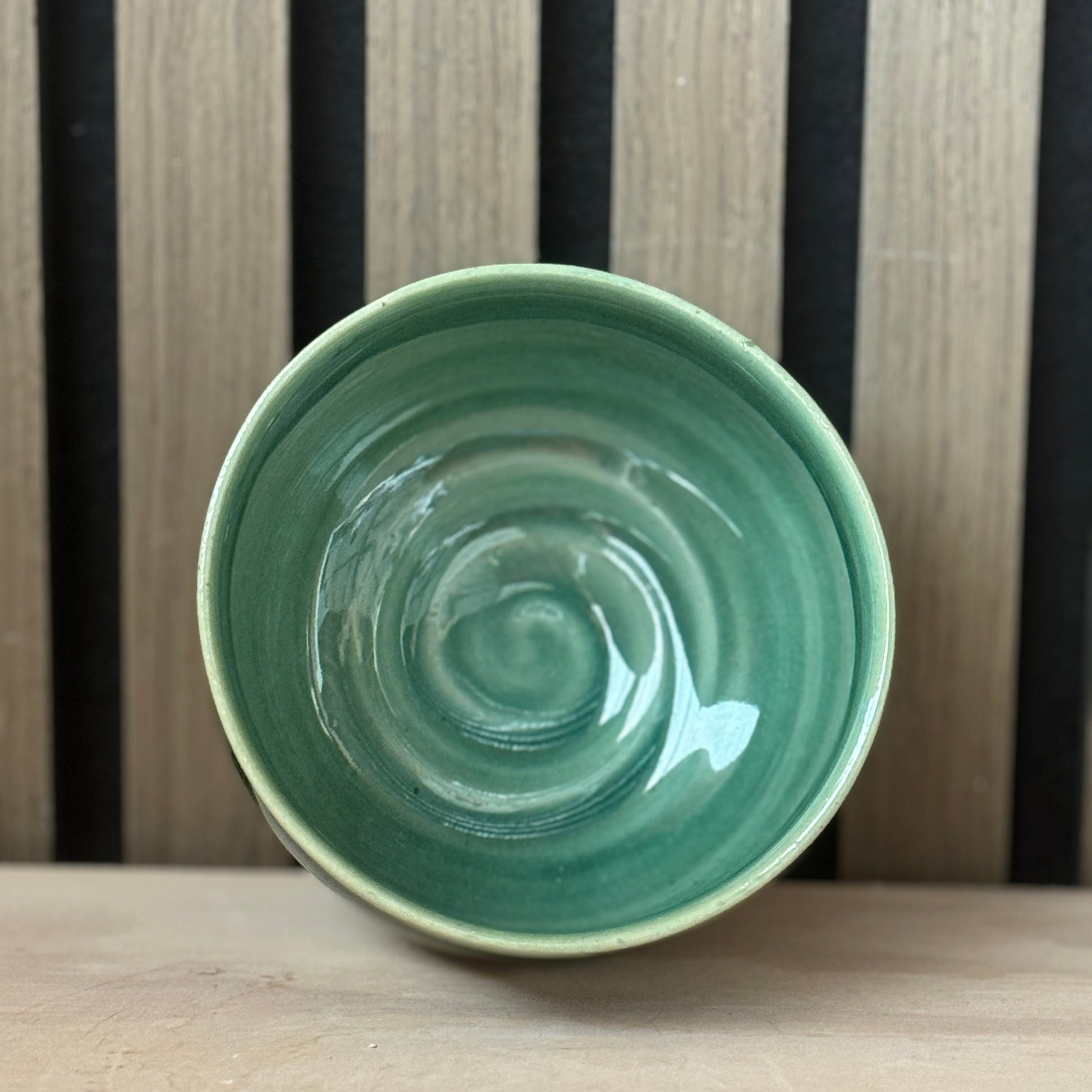 A handmade ceramic bowl with a glossy light green interior and a contrasting beige rim, shown from a top-down perspective on a wooden surface against a striped wooden background.