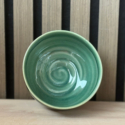 A handmade ceramic bowl with a glossy light green interior and a contrasting beige rim, shown from a top-down perspective on a wooden surface against a striped wooden background.