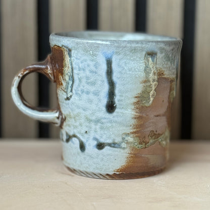 A handmade ceramic mug with a rustic, hand-painted glaze featuring brown, beige, and blue-gray tones, sitting on a light wooden surface.
