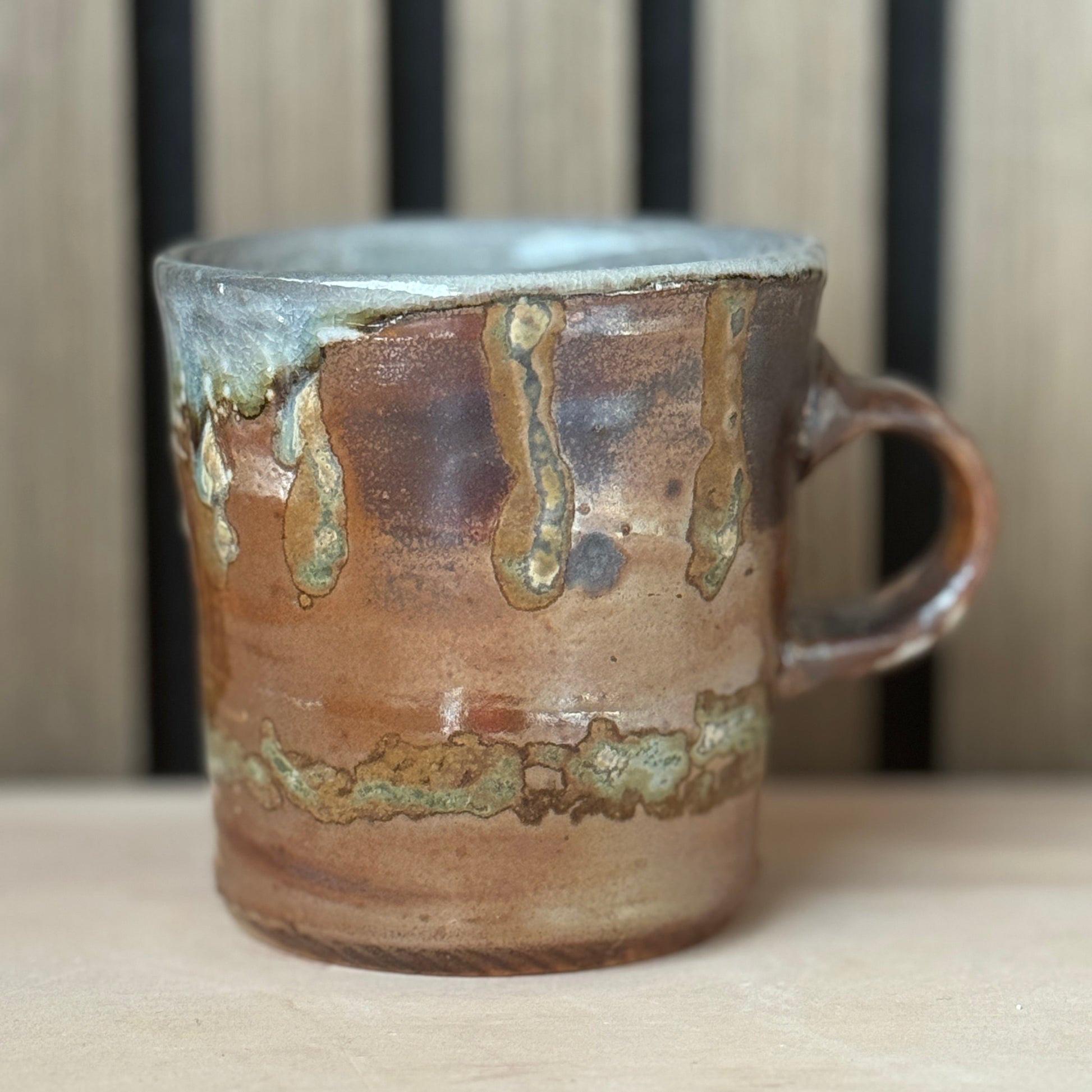 A handmade ceramic mug with a rustic dripping glaze finish, featuring a warm brown and coppery orange body with metallic gold and silver accents, set against a wooden slat background.