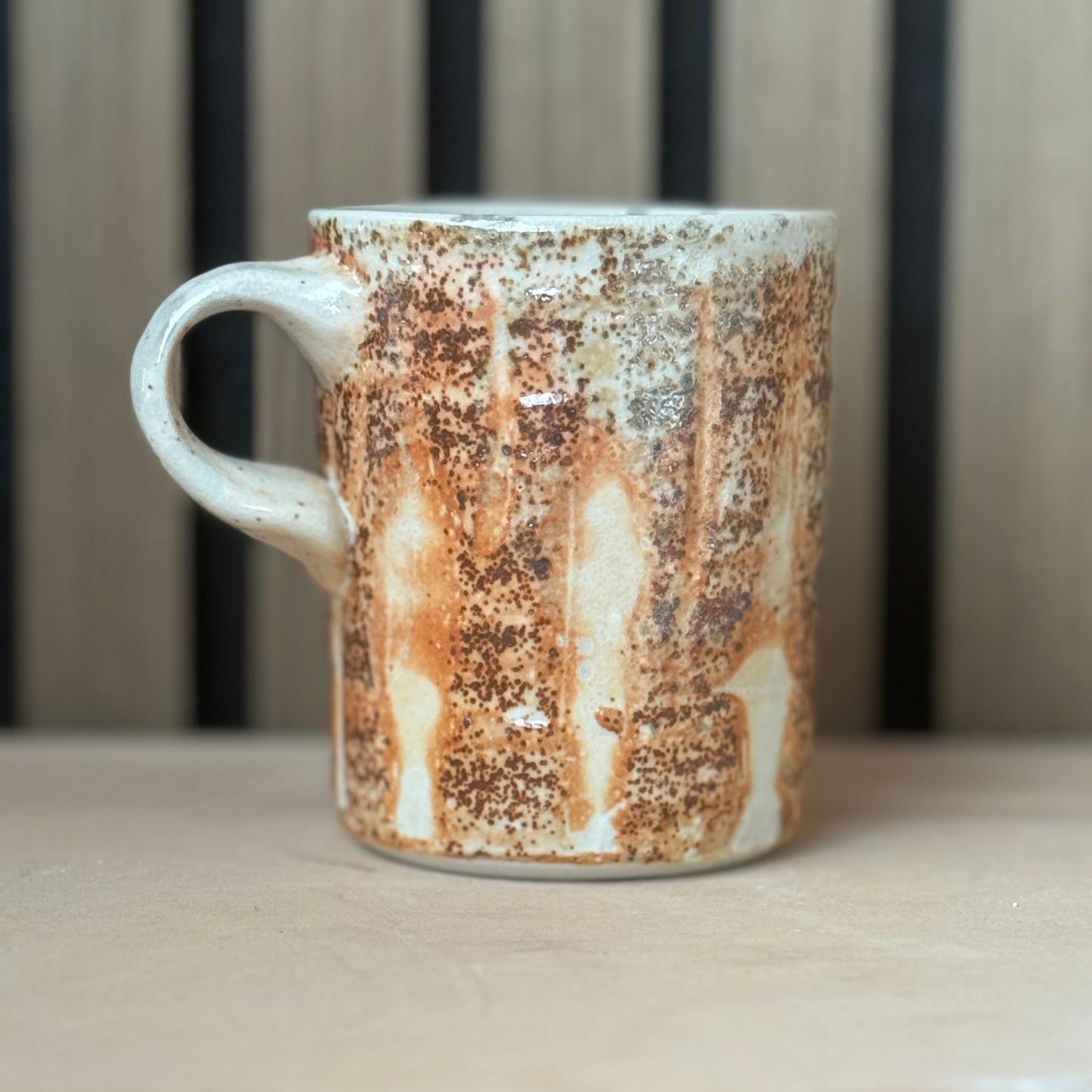 A handmade ceramic mug with a rustic, distressed glaze featuring brown and orange rust-like patterns over a cream base, sitting on a light wooden surface.