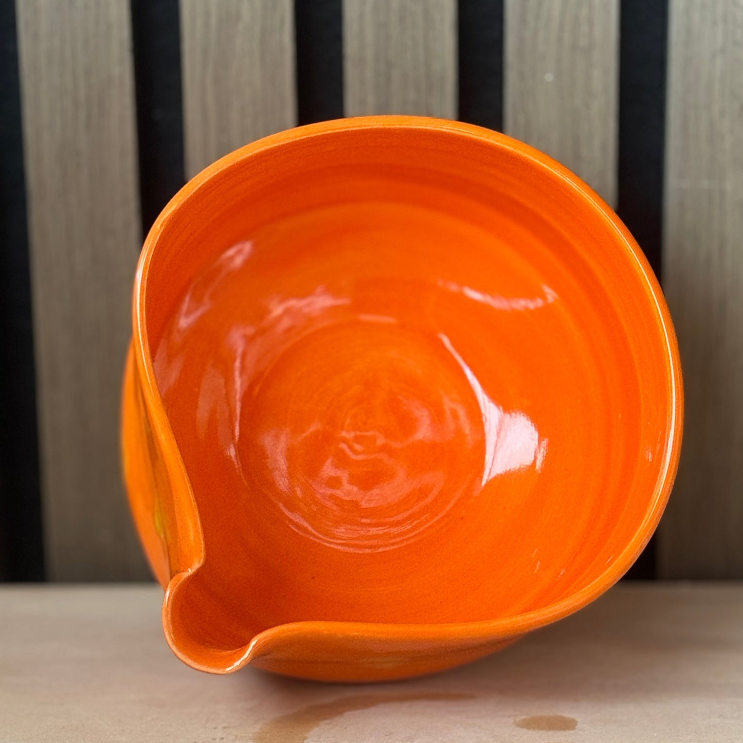 A glossy bright orange ceramic bowl with a small pouring spout, shown from an elevated angle against a background of vertical black and light wood slats.