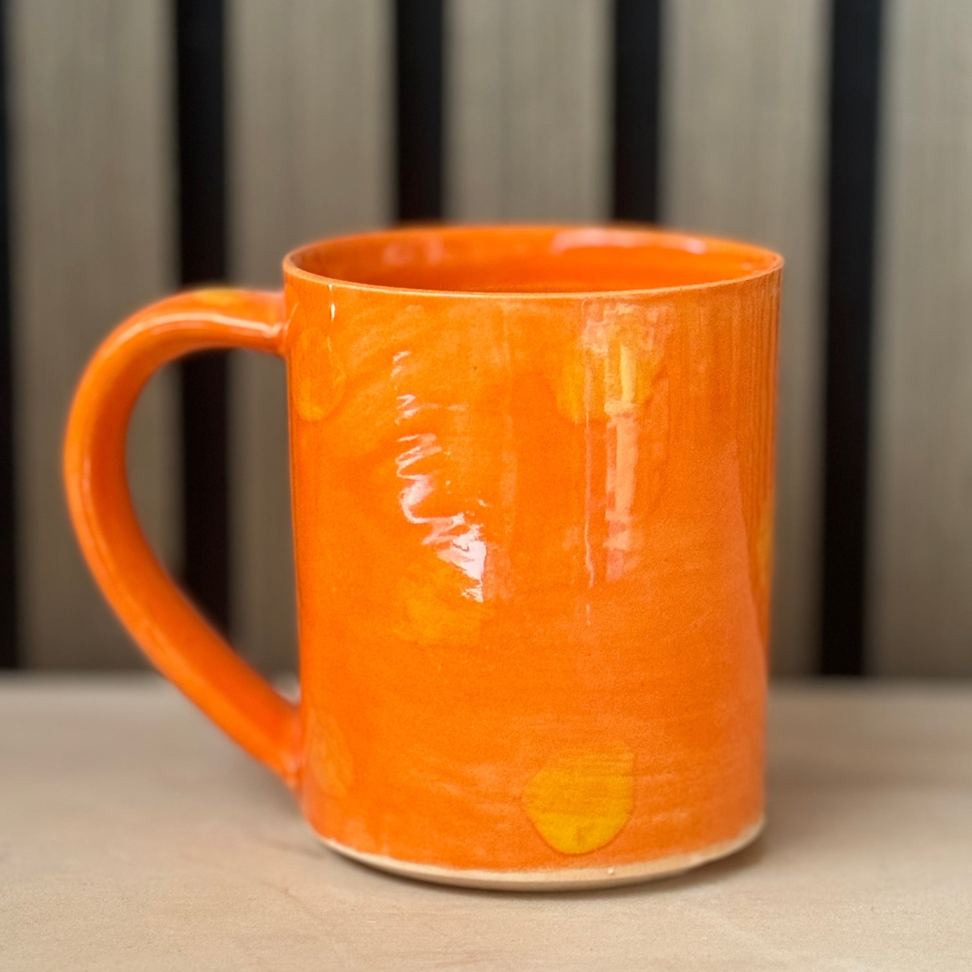 An orange ceramic mug with a glossy finish and subtle yellow hand-painted accents, sitting on a light wooden surface with a blurred background.