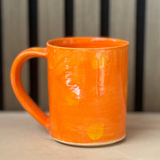 An orange ceramic mug with a glossy finish and subtle yellow hand-painted accents, sitting on a light wooden surface with a blurred background.