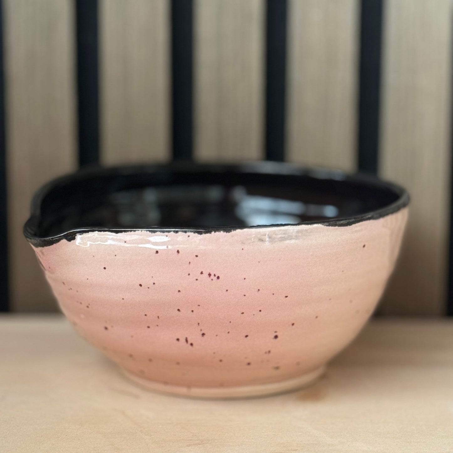 A handmade ceramic bowl with a speckled pink exterior and a glossy black interior, resting on a light wooden surface with a slatted wood background.