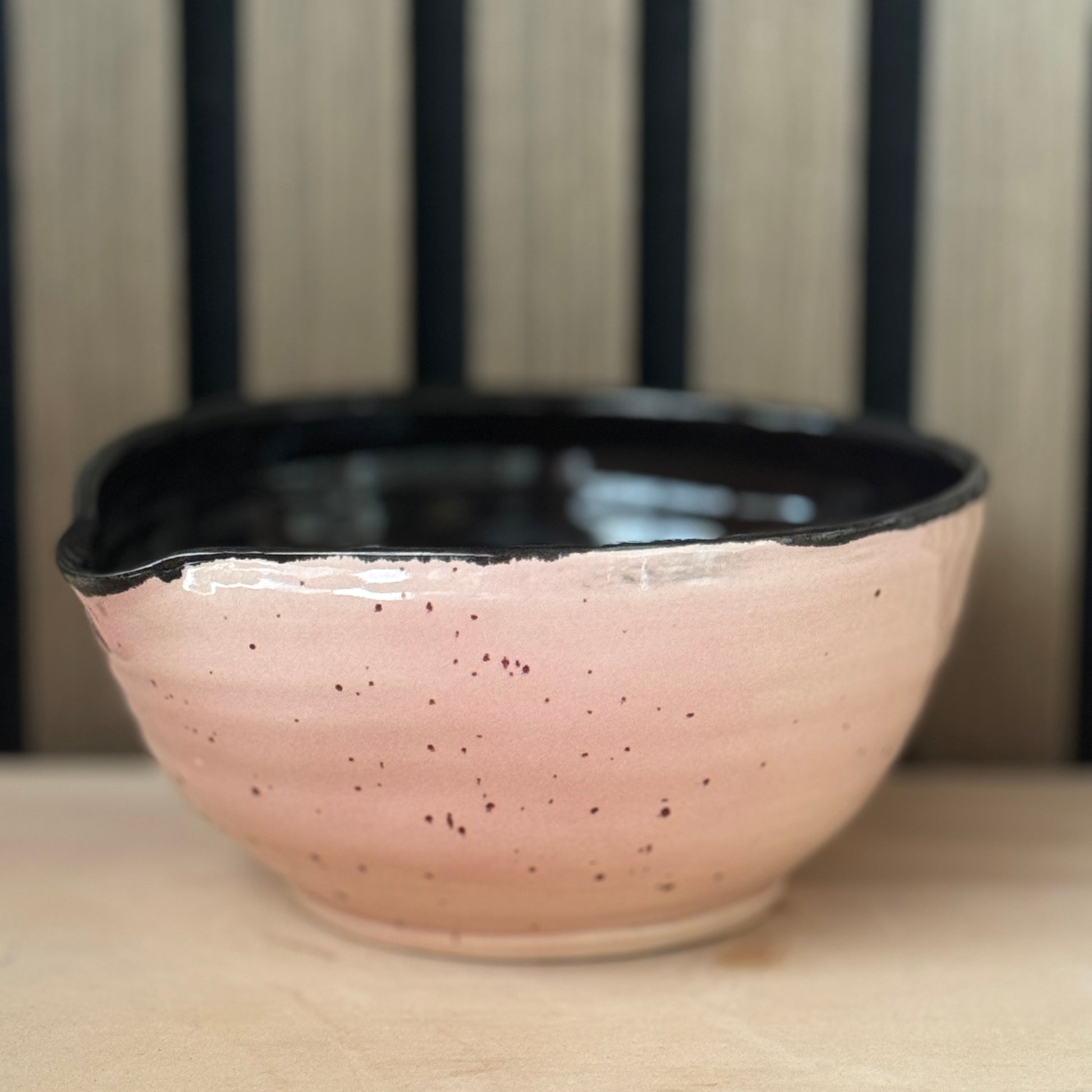 A handmade ceramic bowl with a speckled pink exterior and a glossy black interior, resting on a light wooden surface with a slatted wood background.