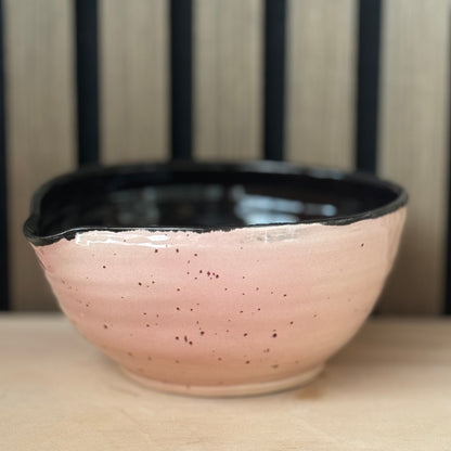 A handmade ceramic bowl with a speckled pink exterior and a glossy black interior, resting on a light wooden surface with a slatted wood background.
