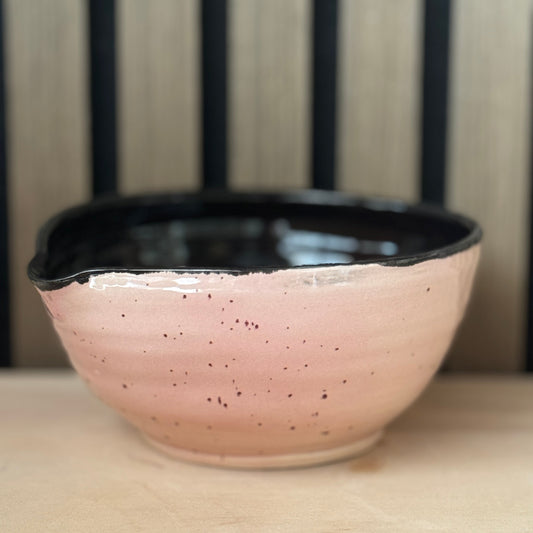 A handmade ceramic bowl with a speckled pink exterior and a glossy black interior, resting on a light wooden surface with a slatted wood background.