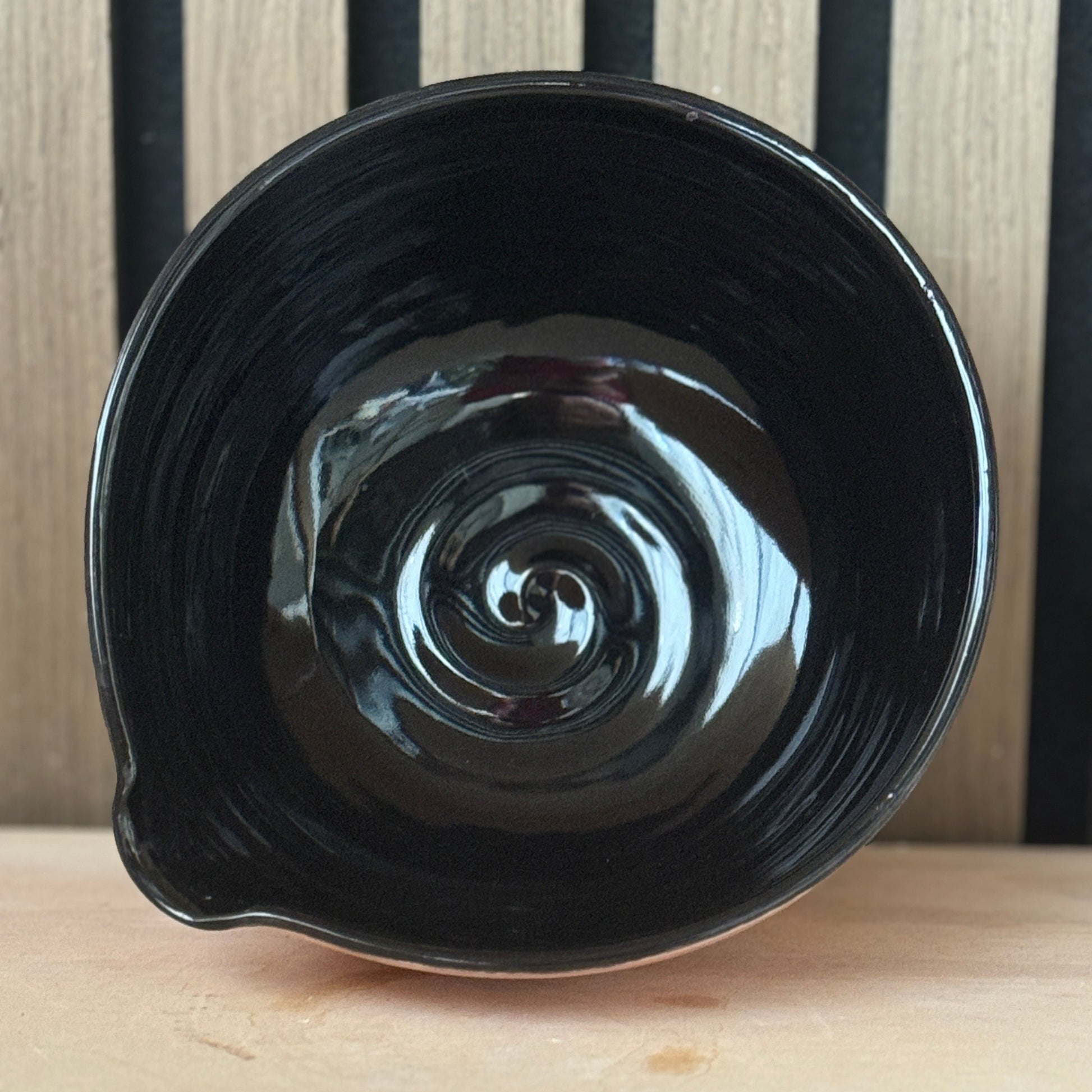 A glossy black ceramic bowl with a spiral pattern on the inside, resting on a light wooden surface against a background of vertical dark wood slats.