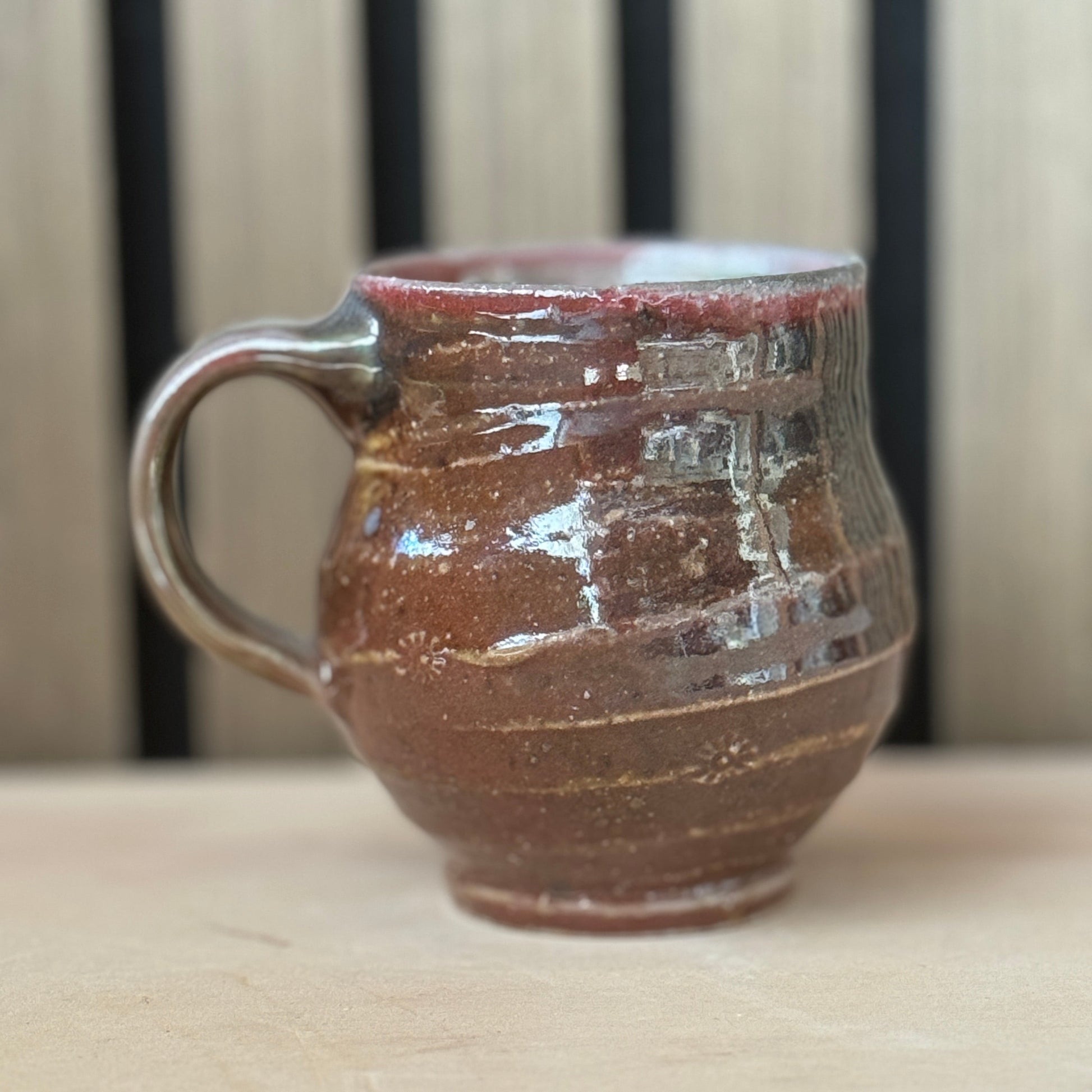 A handmade ceramic mug with a rounded body, a thick handle, and a rustic textured glaze in earthy brown and reddish tones, sitting on a light wooden surface with a blurred wooden slat background.