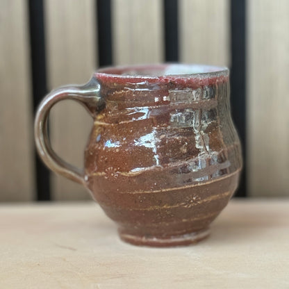 A handmade ceramic mug with a rounded body, a thick handle, and a rustic textured glaze in earthy brown and reddish tones, sitting on a light wooden surface with a blurred wooden slat background.