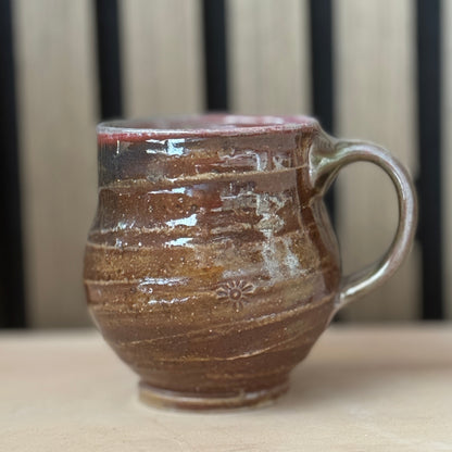 A handmade ceramic mug with a rounded body, a curved handle, and a glossy brown glaze with horizontal textured bands, sitting on a light wooden surface with a blurred background.