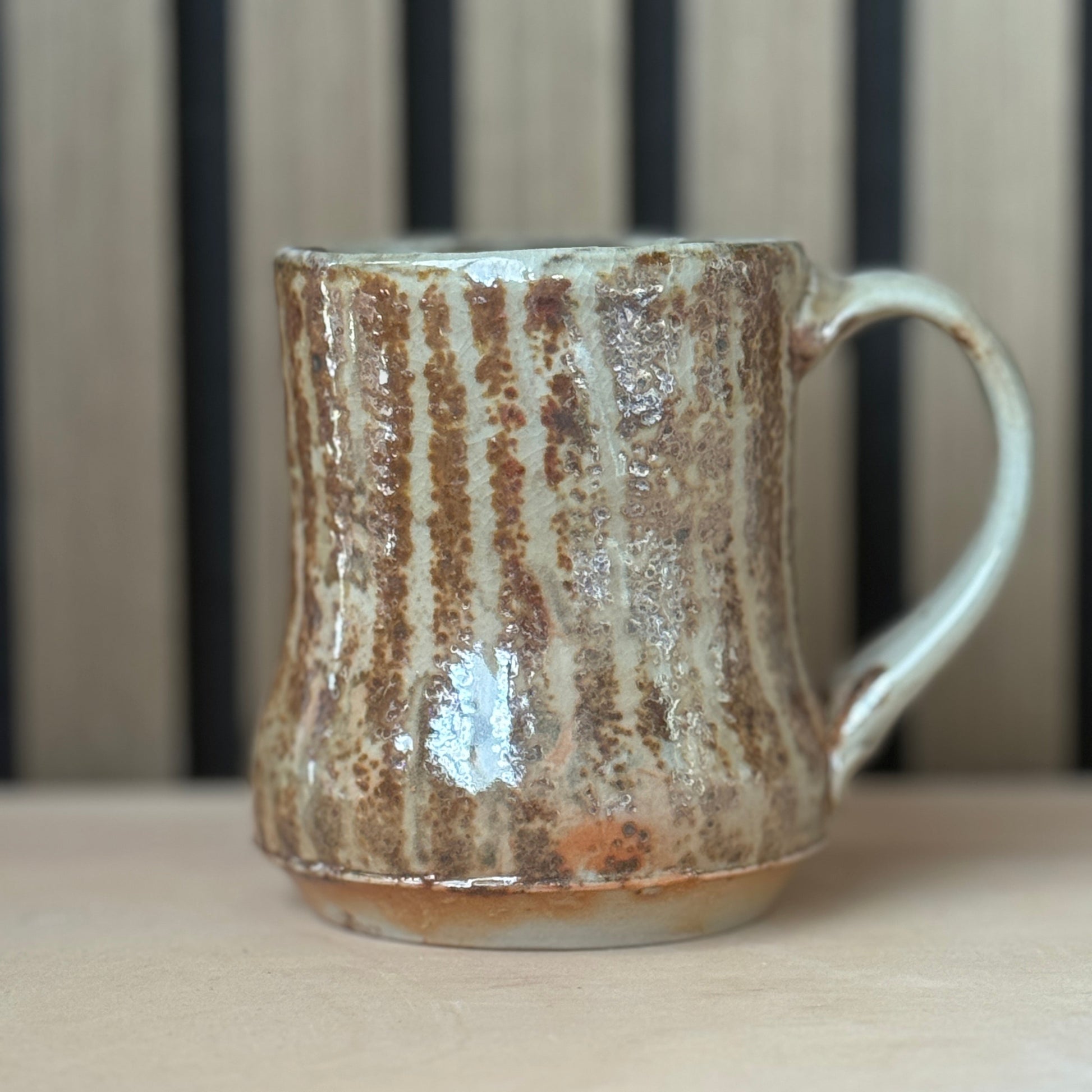 A handmade ceramic mug with a rustic, vertically ribbed glaze in earthy brown and beige tones, sitting on a light wooden surface with a blurred slatted wood background.