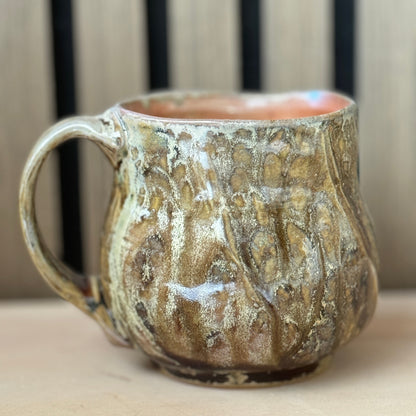 A handmade ceramic mug with a textured, organic shape and a marbled glaze in earthy brown, beige, and gold tones, sitting on a light wooden surface.
