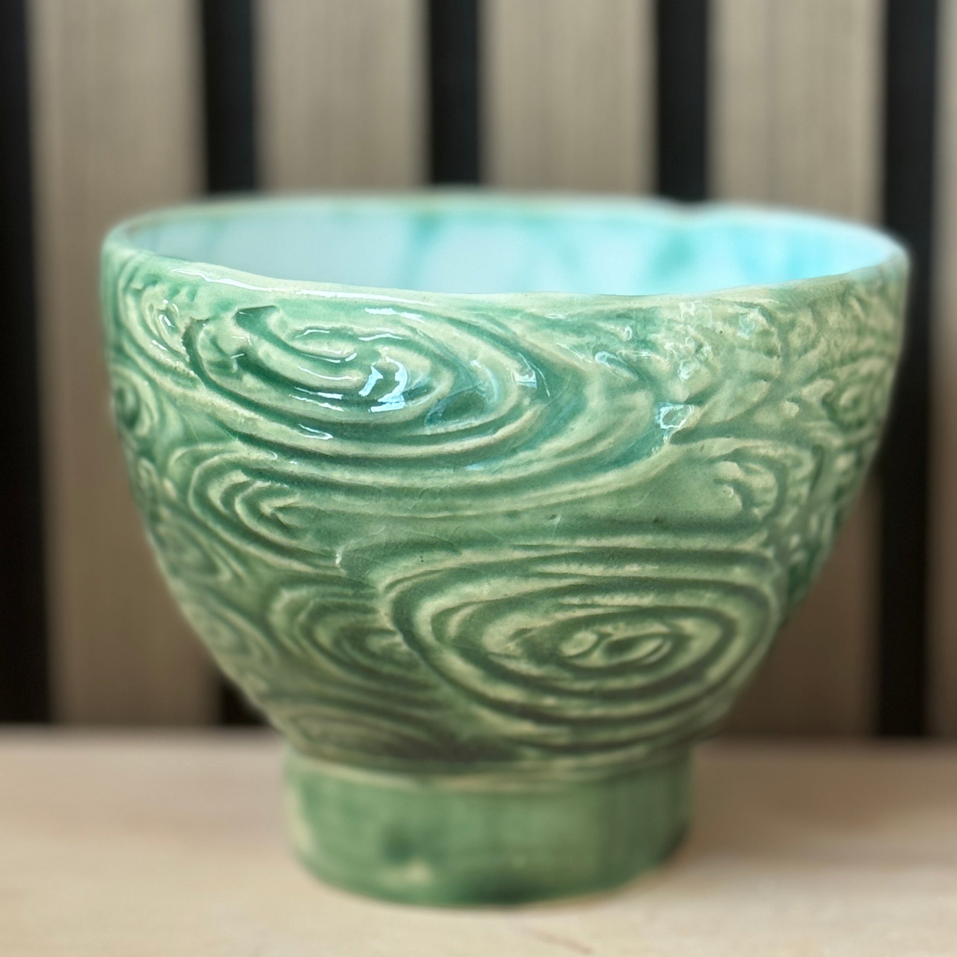 A handmade ceramic bowl with a textured, swirling wave-like pattern in a light green glaze, set against a blurred background of vertical wooden slats.