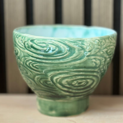 A handmade ceramic bowl with a textured, swirling wave-like pattern in a light green glaze, set against a blurred background of vertical wooden slats.