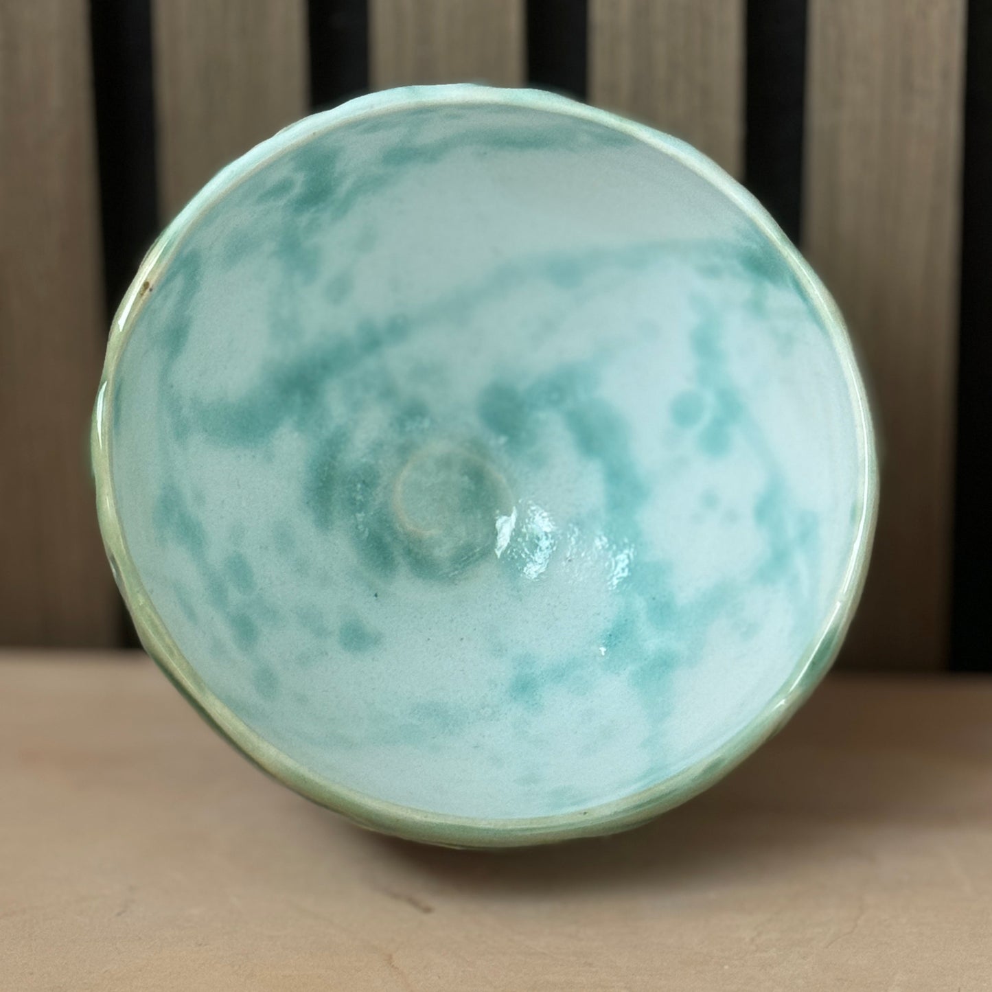 A handmade ceramic bowl with a glossy light green and white marbled glaze, photographed from a top-down angle on a wooden surface.