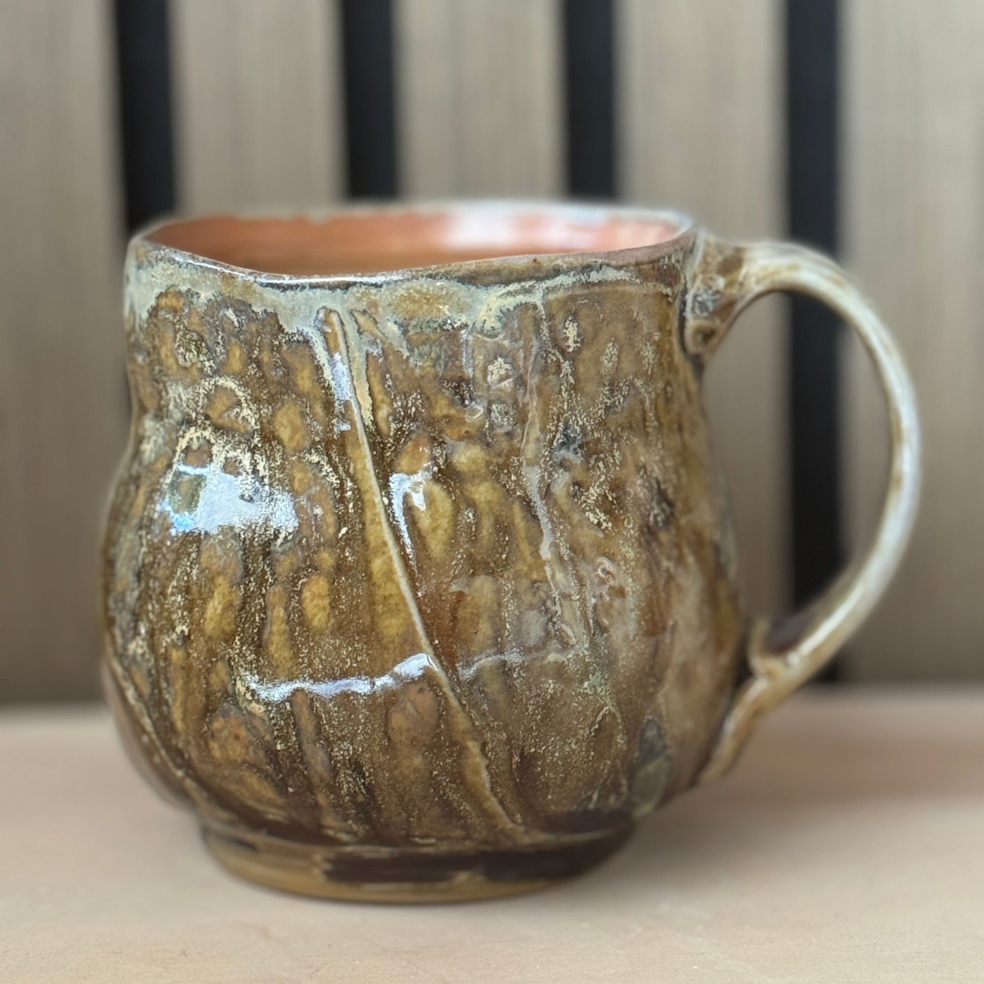 A handmade ceramic mug with a textured, bark-like glaze in earthy brown and gold tones, featuring a coppery orange interior and a curved handle, set against a blurred background of vertical wooden slats.