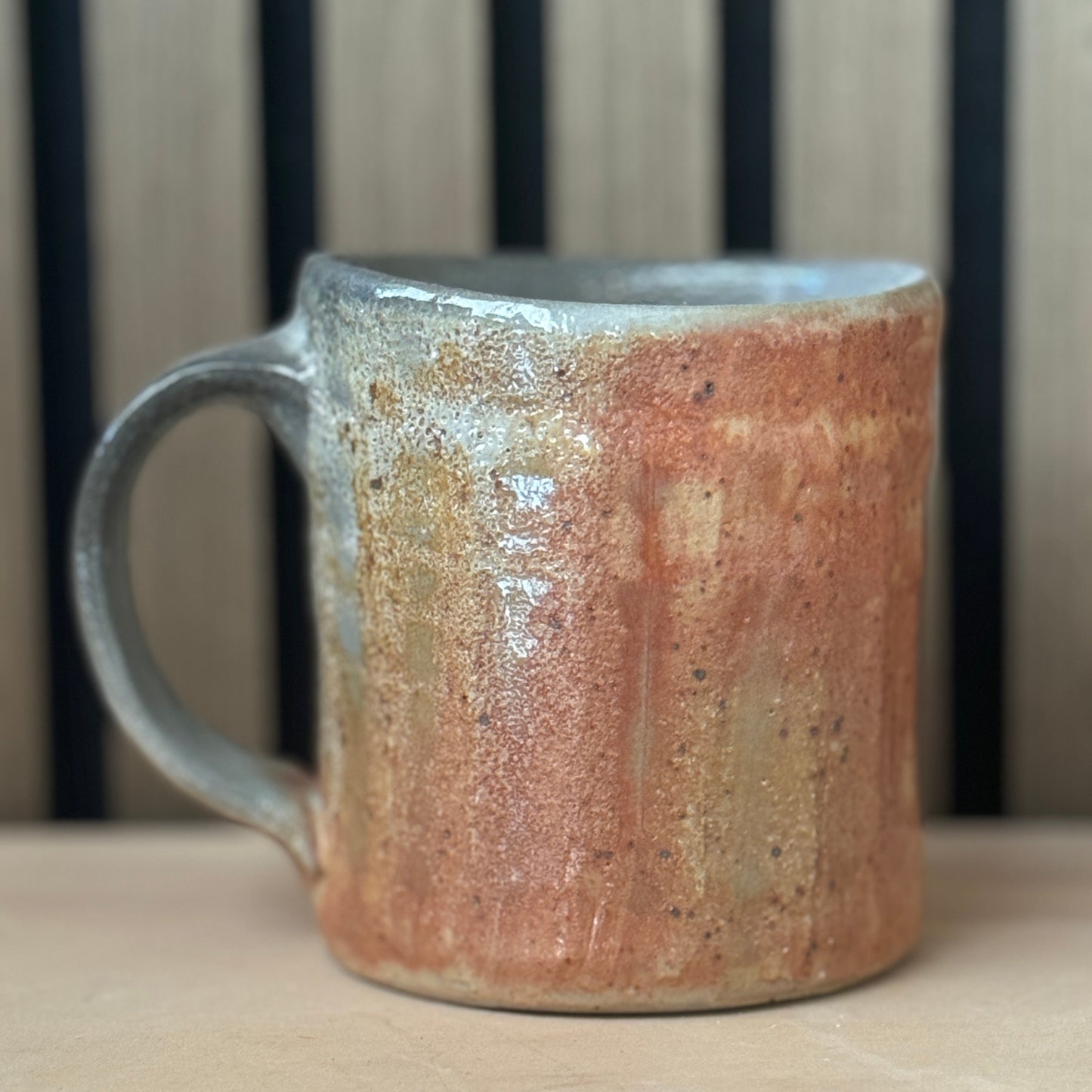 A handmade ceramic mug with a rustic handle, featuring a textured glaze with orange, brown, and gray tones, set against a background of vertical wooden slats.