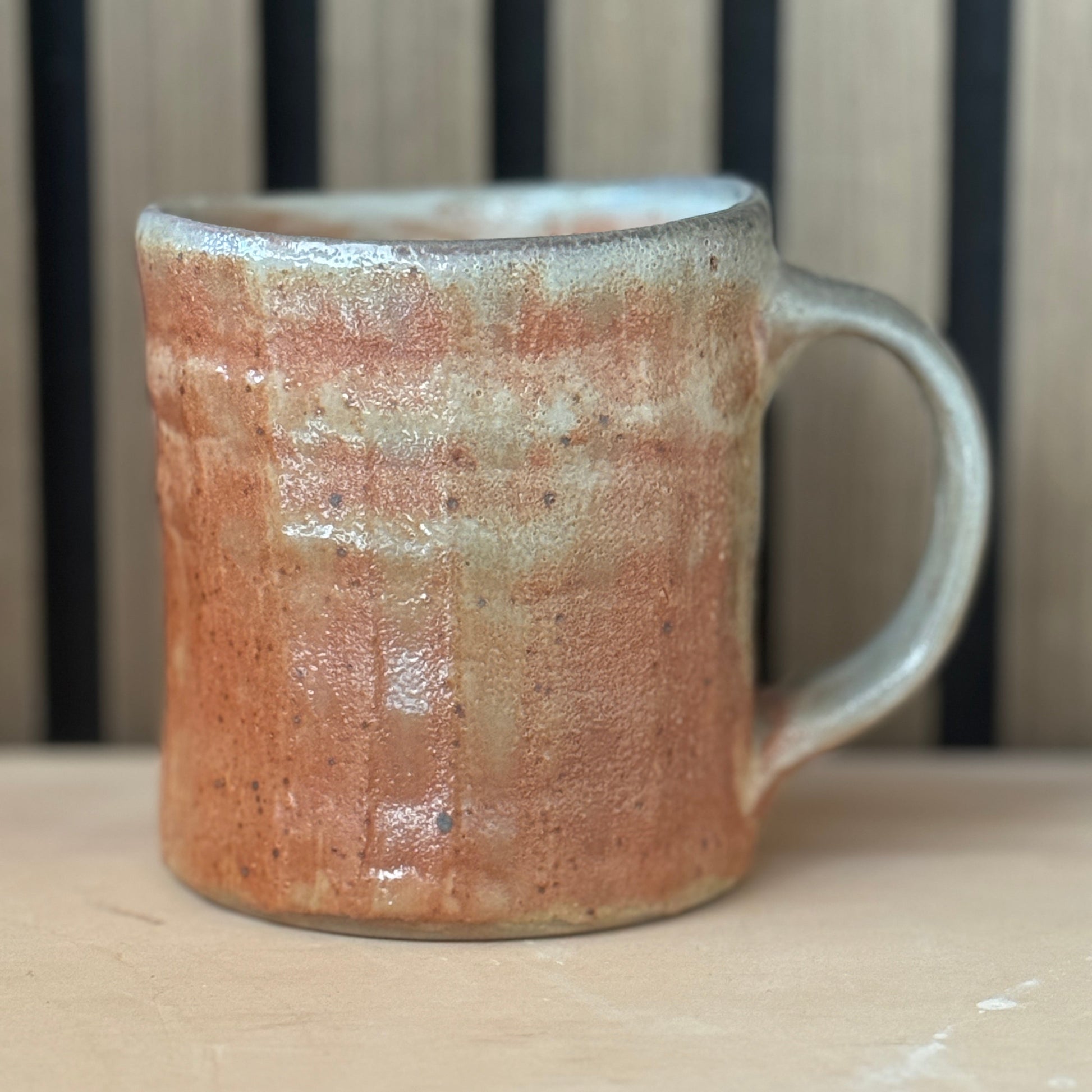 A handmade ceramic mug with a rustic, textured glaze in terracotta orange and light gray, sitting on a light wooden surface with a slatted wood background.
