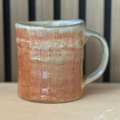 A handmade ceramic mug with a rustic, textured glaze in terracotta orange and light gray, sitting on a light wooden surface with a slatted wood background.