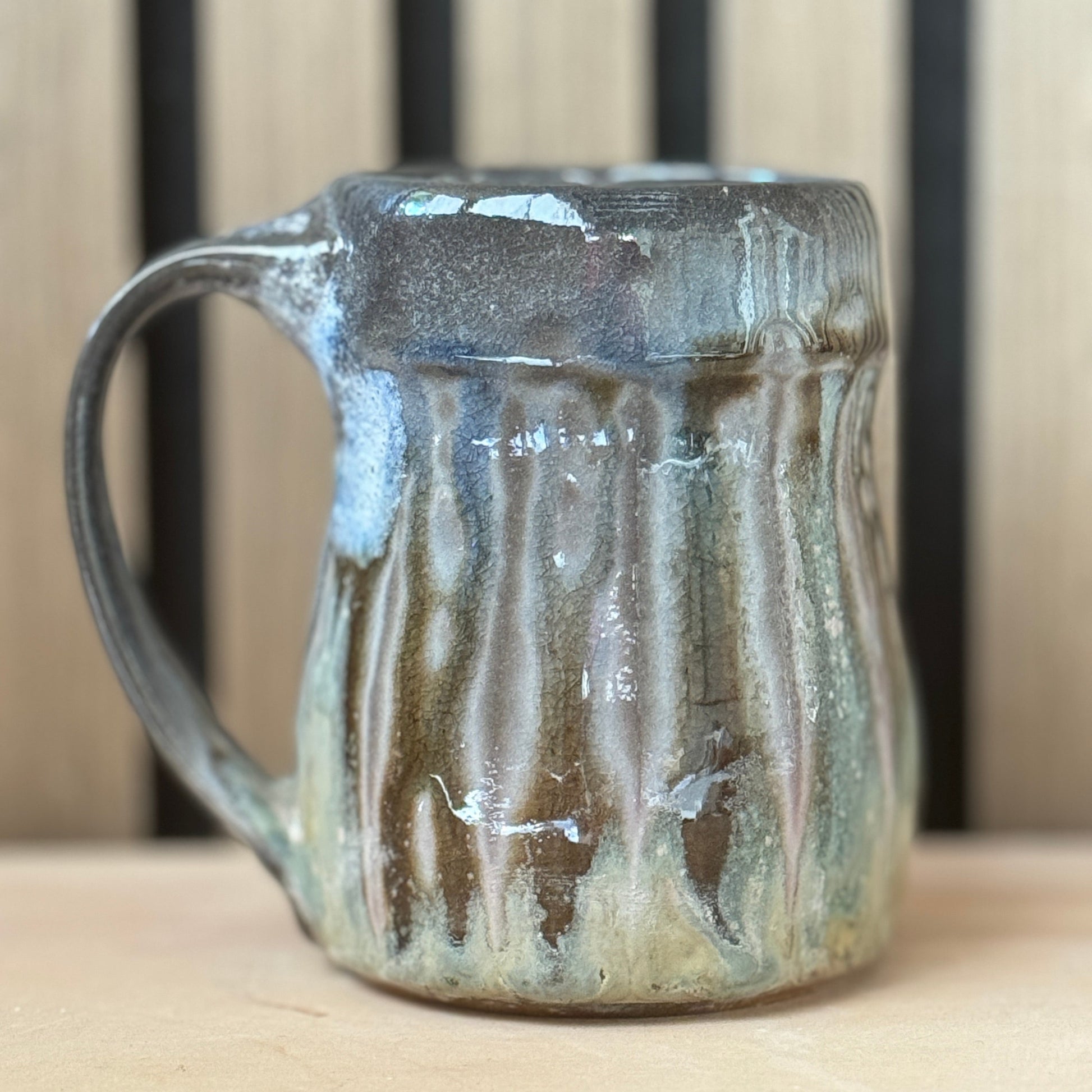 A handmade ceramic mug with a rustic handle, featuring a textured glaze with vertical drips of brown, gray, and light blue-green, set against a wooden slat background.