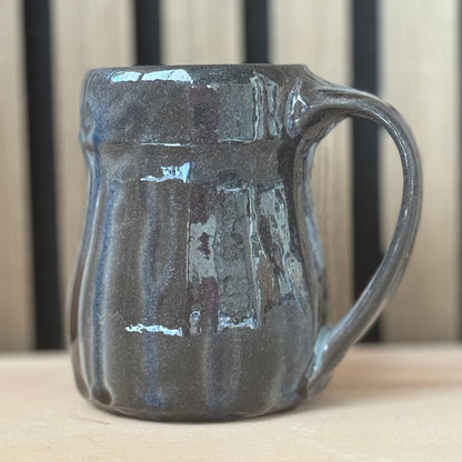 A handmade ceramic mug with a dark charcoal gray and blue-gray glaze, featuring a ribbed texture and a curved handle, set on a light wooden surface with a blurred wooden slat background.