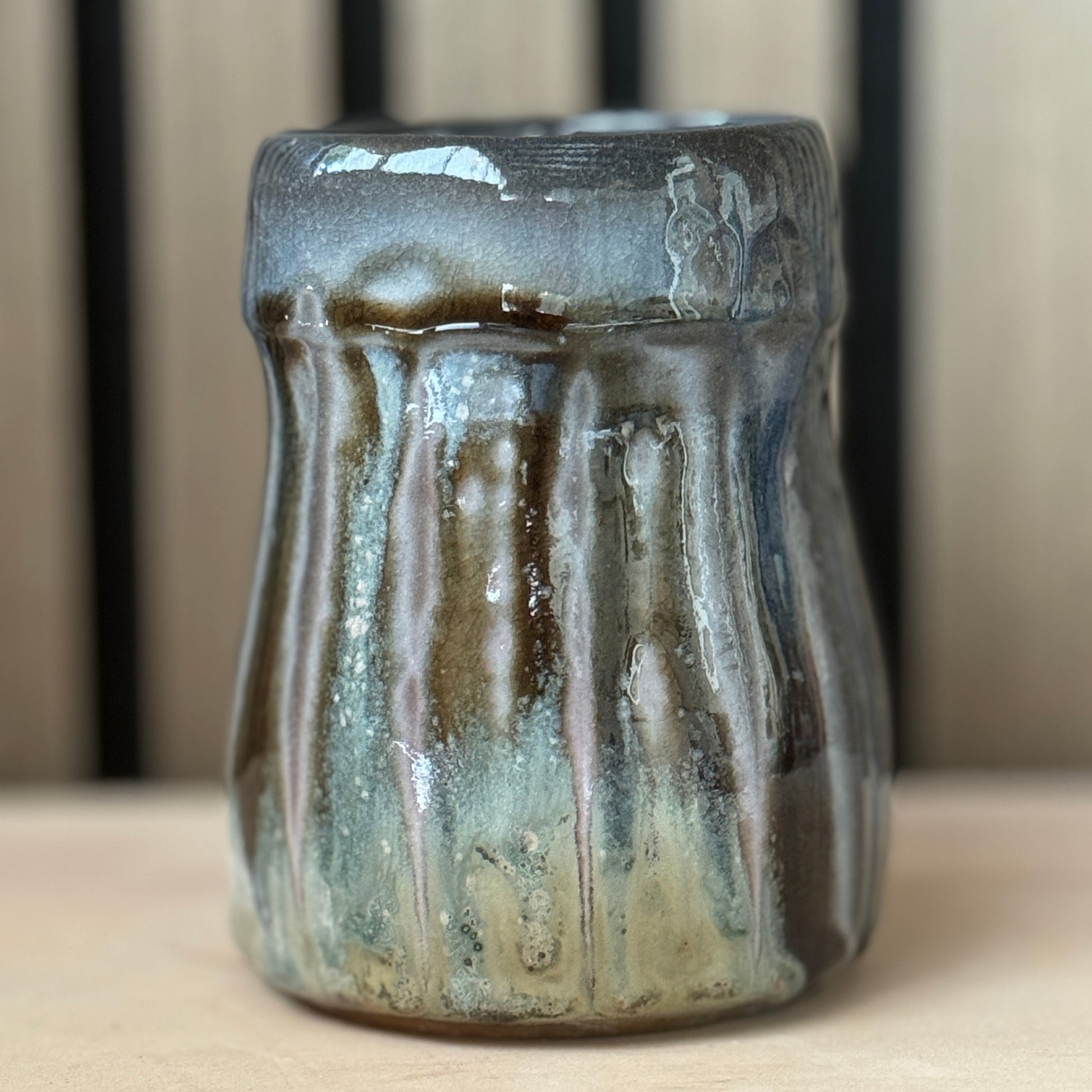 A handmade ceramic cup with a textured, ribbed exterior and a glossy, dripping glaze in shades of brown, gray, and blue-green, set against a neutral wooden background.