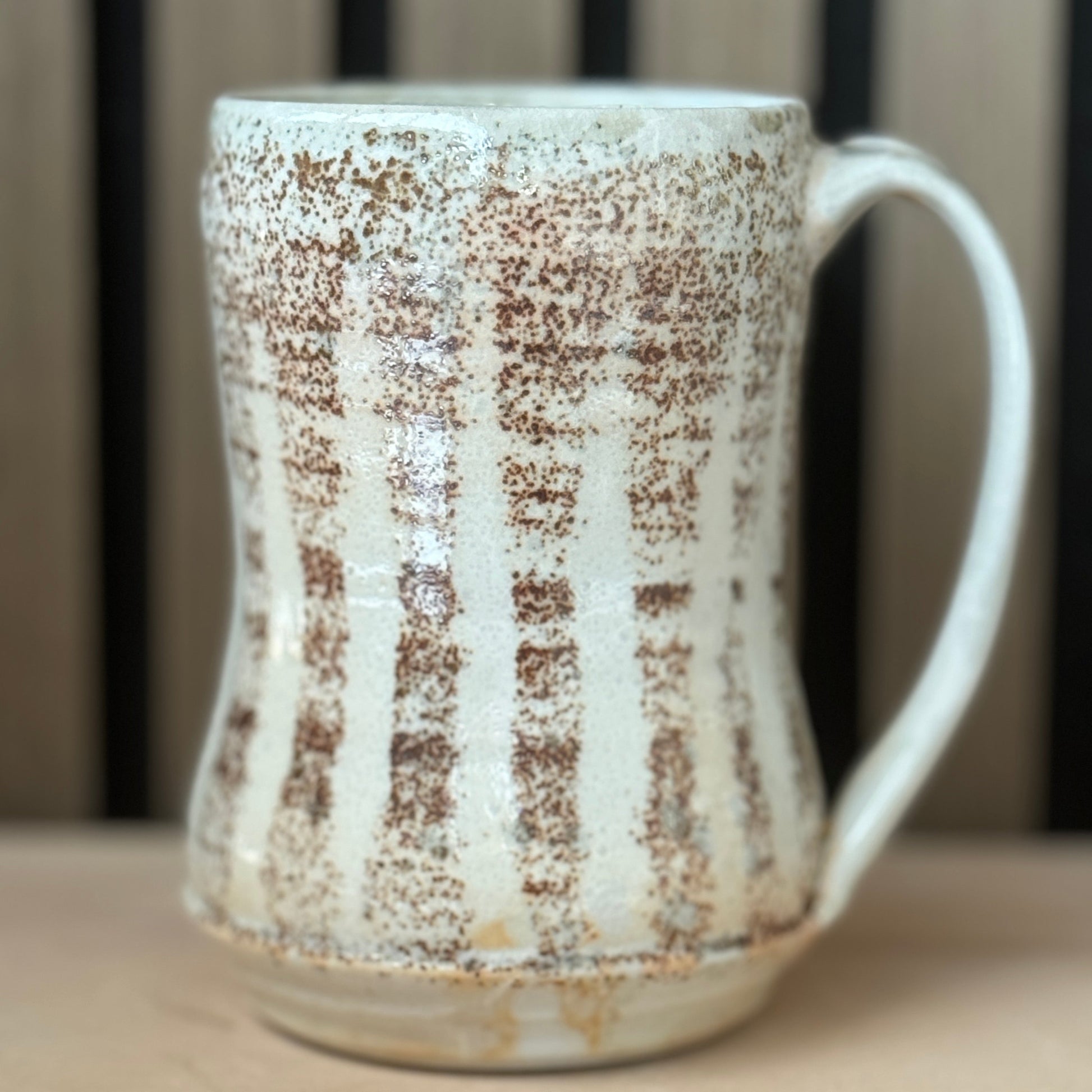 A handmade ceramic mug with a textured, speckled glaze in off-white and brown, featuring a ribbed body and a curved handle, set against a blurred background.