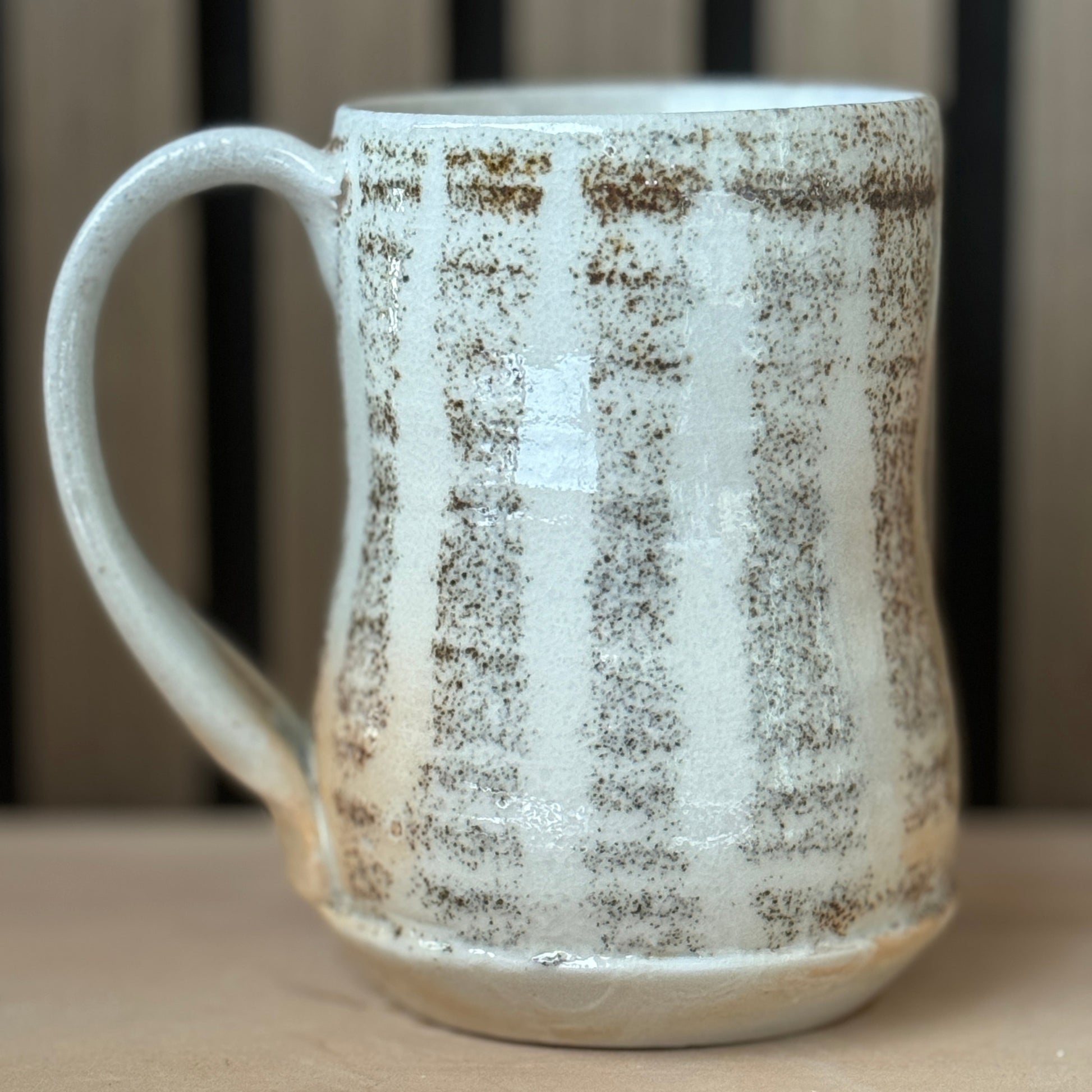 A handmade ceramic mug with a textured, speckled glaze in off-white and brown, featuring a curved handle and a rustic finish, set on a wooden surface.