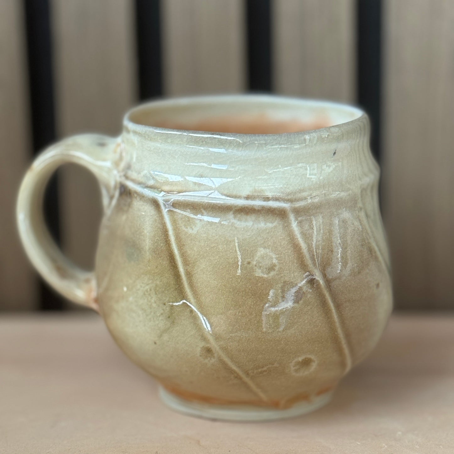 A handmade ceramic mug with a rounded body, a side handle, and a glossy glaze in warm beige and cream tones, set against a blurred background of vertical wooden slats.