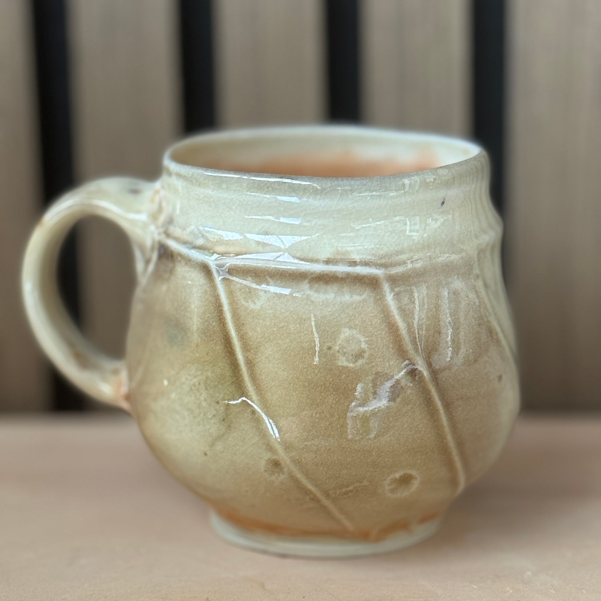 A handmade ceramic mug with a rounded body, a side handle, and a glossy glaze in warm beige and cream tones, set against a blurred background of vertical wooden slats.