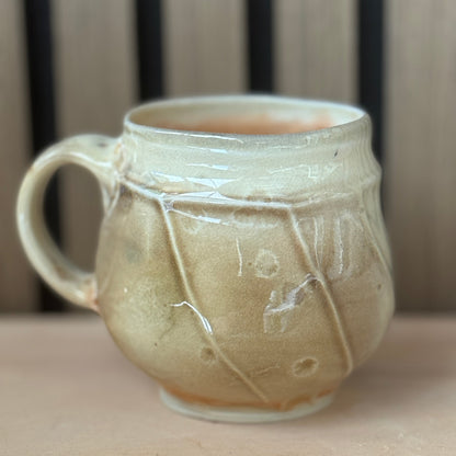 A handmade ceramic mug with a rounded body, a side handle, and a glossy glaze in warm beige and cream tones, set against a blurred background of vertical wooden slats.