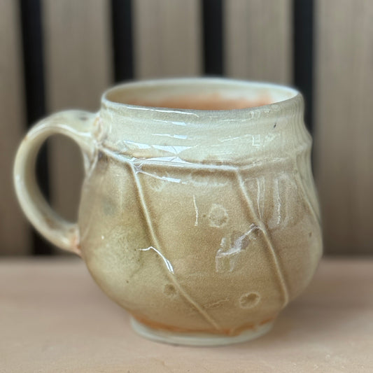 A handmade ceramic mug with a rounded body, a side handle, and a glossy glaze in warm beige and cream tones, set against a blurred background of vertical wooden slats.