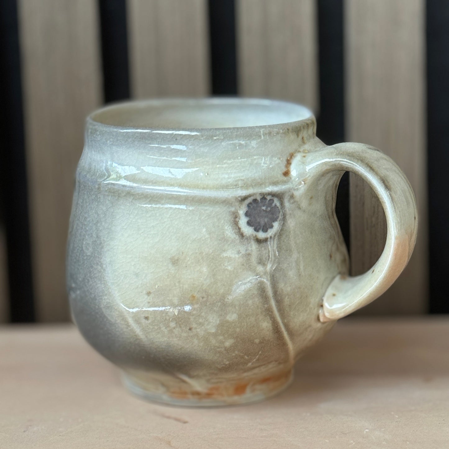 A handmade ceramic mug with a rounded body, a thick handle, and a rustic, textured glaze in earthy beige and gray tones, resting on a wooden surface.