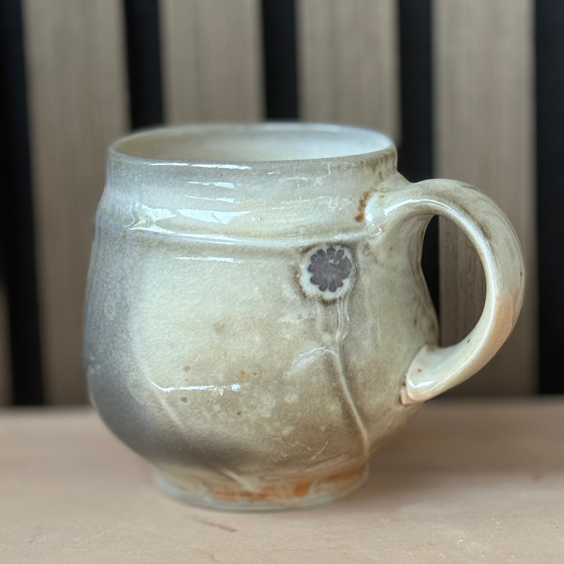 A handmade ceramic mug with a rounded body, a thick handle, and a rustic, textured glaze in earthy beige and gray tones, resting on a wooden surface.