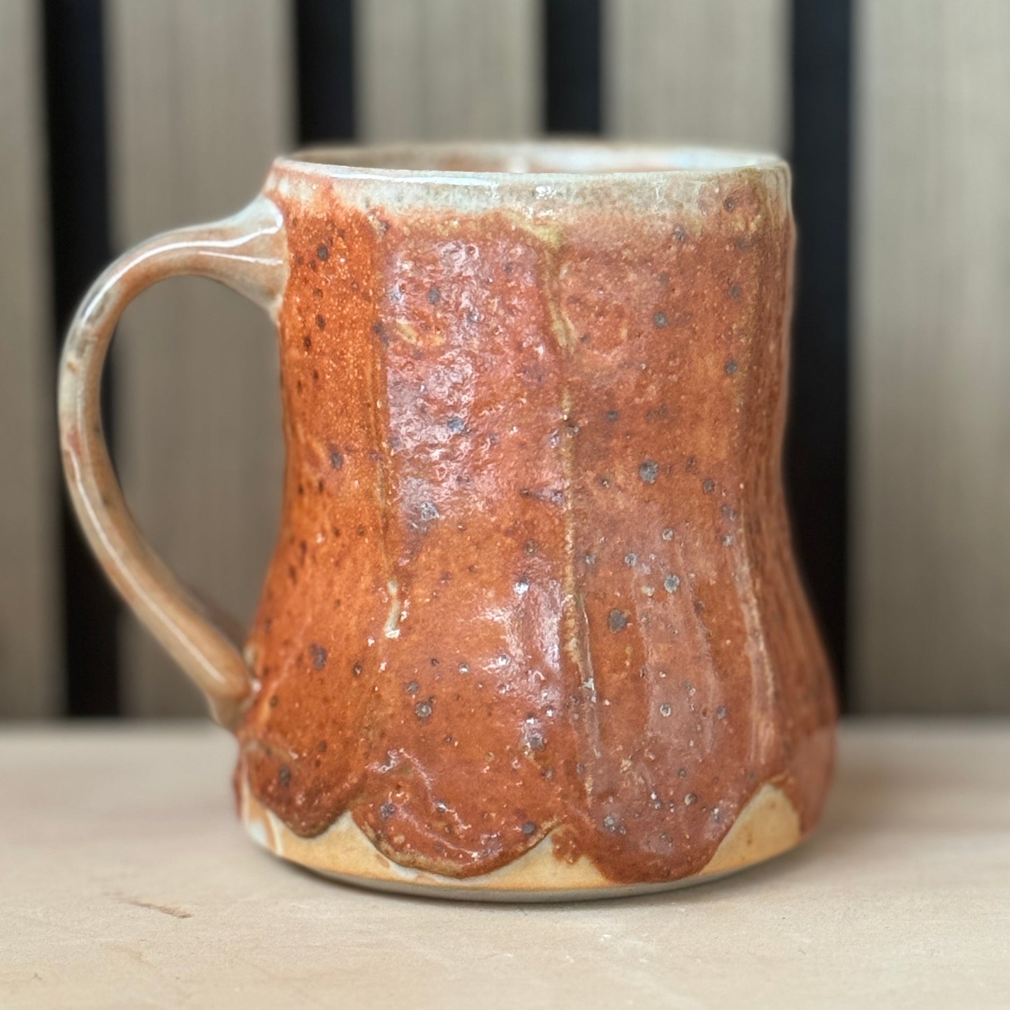 A handmade ceramic mug with a rustic, textured glaze in terracotta brown and orange, featuring a dripping finish and a light beige base, set against a blurred background of vertical wooden slats.