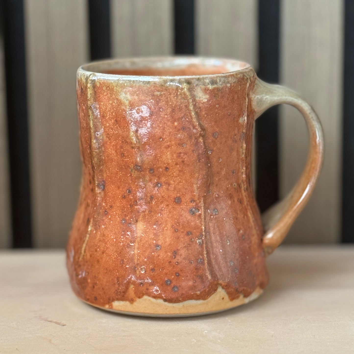 A handmade ceramic mug with a rustic, textured glaze in terracotta orange and brown, featuring a thick handle and a speckled finish, set against a neutral background.