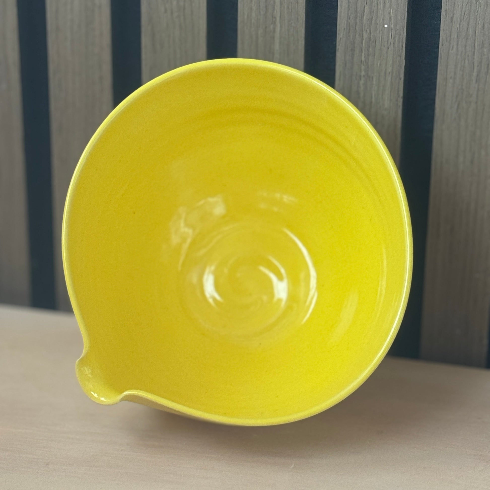 A bright yellow ceramic bowl with a small pouring spout, shown from a high angle on a light wooden surface against a dark wood slatted background.