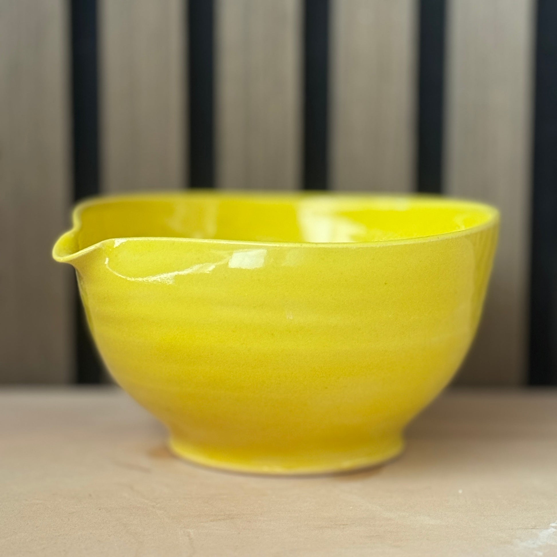 A bright yellow ceramic bowl with a small pouring spout, resting on a light beige surface against a blurred background of vertical dark stripes.