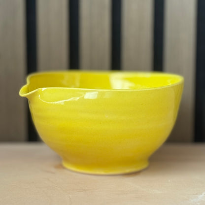 A bright yellow ceramic bowl with a small pouring spout, resting on a light beige surface against a blurred background of vertical dark stripes.